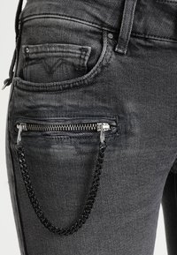 Acercamiento de unos jeans de mezclilla grises que muestran un bolsillo con cremallera y un accesorio de cadena negra adjunto en el lado derecho.