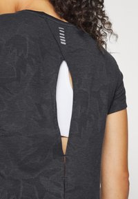 Svart träningsskjorta med ett strukturerat mönster, med en skärning bak och korta ärmar. En vit lager under är synlig genom skärningen.