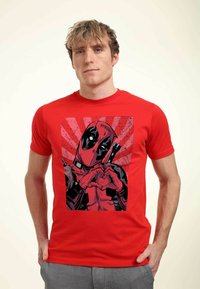 Červené bavlněné tričko s grafickým potiskem postavy, která tvoří tvar srdce, s černobílými detaily a tečkovaným pozadím.