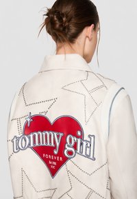 Jachetă din piele albă cu ținte argintii și desene cu stele. Pe spate are o inimă mare roșie cu „tommy girl” și „FOREVER” brodate în albastru.