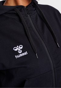 Sudadera con cremallera negra hecha de tela suave, que presenta un logo de abeja blanca bordado en el pecho y cordones texturizados en la capucha.