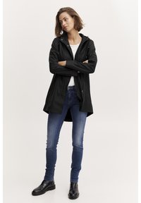 Svart vattentät jacka med huva, dragkedja fram, och justerbara snoddar, kombinerad med tajta blå jeans och ankelstövlar.