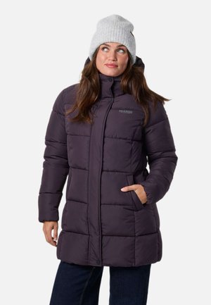 Une femme présente un manteau matelassé couleur prune foncé et un bonnet blanc en maille, une main dans la poche, devant un fond uni.
