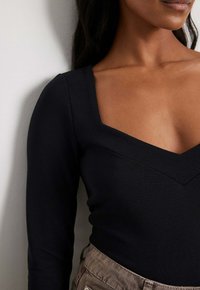 Haut à manches longues noir à côtes, présentant un large col en V et une silhouette ajustée. La texture est lisse avec des détails subtils au niveau des coutures.