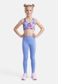 Νεαρή κοπέλα με πλεξίδες σε διπλούς κότσους, φορώντας αθλητικό τοπ με λουλούδια, ανοιχτό μπλε κολάν και λευκά-ροζ αθλητικά παπούτσια, στέκεται με αυτοπεποίθηση.