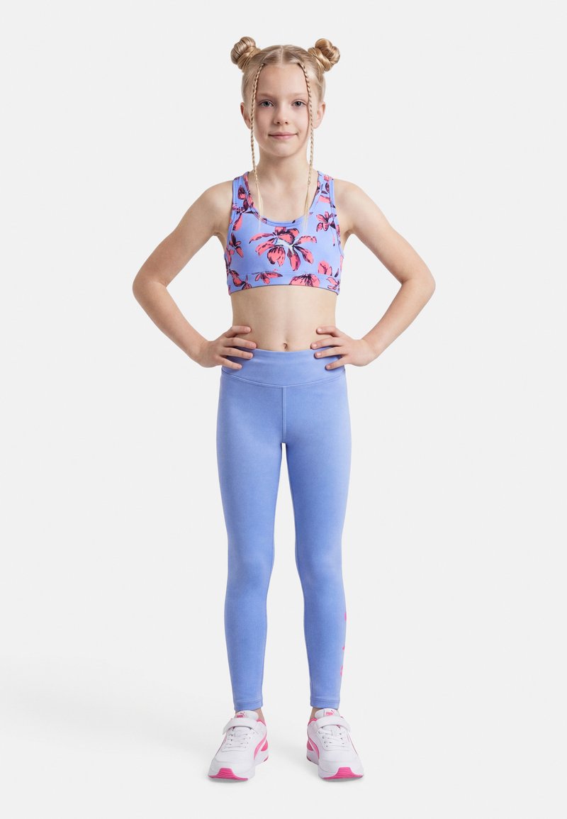 Νεαρή κοπέλα με πλεξίδες σε διπλούς κότσους, φορώντας αθλητικό τοπ με λουλούδια, ανοιχτό μπλε κολάν και λευκά-ροζ αθλητικά παπούτσια, στέκεται με αυτοπεποίθηση.