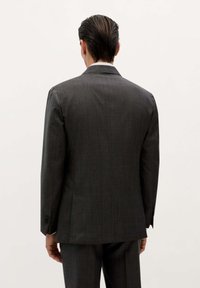 Hombre con el cabello liso y oscuro, vistiendo un traje gris oscuro, de pie de espaldas contra un fondo claro y uniforme.
