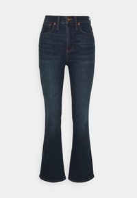 Jeans de mezclilla de tiro alto en azul oscuro con ligero desgastado, cinco bolsillos, cierre de botón y cremallera, y piernas acampanadas sobre fondo blanco.