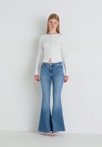 Top blanco de manga larga con ribetes y cierre de botones, combinado con jeans acampanados de color azul claro. Líneas limpias y diseño ajustado con texturas suaves.