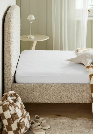 Beiges, strukturiertes Bett mit weißer Matratze und Kissen, runder Beistelltisch mit weißer Lampe, kariertem Rucksack, beigen Schuhen und transparenten Vorhängen im Hintergrund.