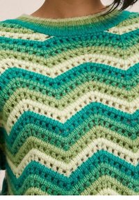 Close-up van een gebreide trui met groene, turquoise en crèmekleurige zigzagstrepen en een ronde hals, gedragen door een persoon.