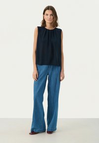 Navy blaues ärmelloses Top mit Falten, kombiniert mit weiten blauen Denim-Hosen und bordeauxfarbenen Schuhen, vor einem neutralen Hintergrund.