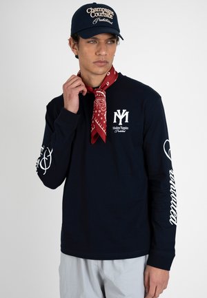 Mężczyzna w granatowej koszuli z długim rękawem z białymi nadrukami, czerwonym wzorzystym szaliku i granatowej czapce z napisem „Champagne Courtside Prohibited”.