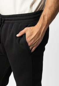 Svarta mjukisbyxor med dragsko i midjan, mjuk tygtextur och sidofickor, visade med en hand vilande i fickan.