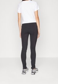 Schwarze, schmal geschnittene Hose mit glatter Textur, kombiniert mit einem weißen T-Shirt mit kurzen Ärmeln und grauen Sneakers, von hinten betrachtet.