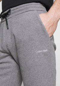 Pantalon de jogging gris en tissu doux, doté d'une taille élastique avec cordon de serrage et d'un logo Calvin Klein blanc sur la cuisse.