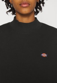 Sudadera negra de cuello alto, hecha de una tela suave, con un pequeño parche rectangular del logo de Dickies en el lado izquierdo del pecho. Textura suave.