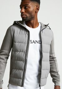 Giacca imbottita grigia con cappuccio e maniche a contrasto. Presenta una zip frontale e tasche laterali. Indossata sopra una t-shirt bianca con il testo "SAND".