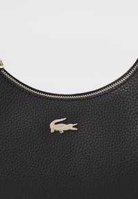 Sac en cuir noir texturé avec une fermeture éclair argentée et un petit logo crocodile argenté centré sous la fermeture éclair.
