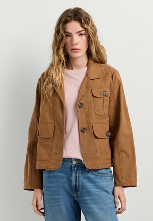 Junge Frau mit welligem blondem Haar trägt eine braune, geknöpfte Jacke über einem rosa Hemd und blauen Jeans, steht vor einem schlichten hellen Hintergrund.