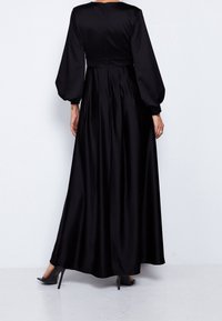 Kvinna i en golvlång svart klänning med långa puffärmar och åtsittande midja, står med ryggen mot kameran, bär svarta högklackade skor.