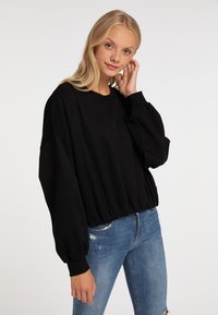 Giovane donna con lunghi capelli biondi indossa una felpa nera oversized e jeans strappati blu, toccando i capelli con una mano su sfondo bianco.