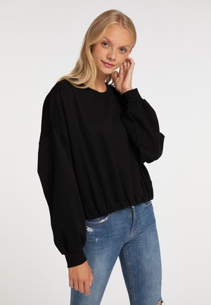 Jonge vrouw met lang blond haar draagt een losse zwarte sweatshirt en blauwe ripped jeans, terwijl ze met één hand haar haar aanraakt tegen een witte achtergrond.