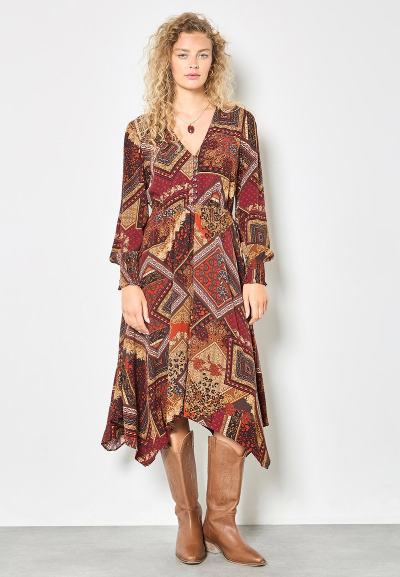 Langes, fließendes Kleid mit einem geometrischen Muster in Rot-, Braun- und goldenen Tönen. Mit einem tiefen V-Ausschnitt und langen Ärmeln, kombiniert mit braunen Cowboystiefeln.