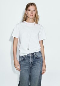 T-shirt bianco in cotone con maniche corte, abbinato a jeans in denim blu a vestibilità ampia. Presenta un collana con pendente in argento come accessorio.