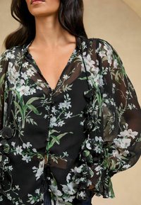 Schwarze, durchsichtige Bluse mit Blumenmuster, mit lockeren Ärmeln und durchgehender Knopfleiste. Akzente beinhalten Stoffblumen entlang der Kanten.
