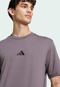 T-shirt grigio a maniche corte con collo rotondo. Presenta il logo Adidas nero sul petto. Il tessuto sembra liscio e leggero.