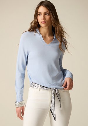 Ung kvinde med langt hår iført en lyseblå sweater og hvide bukser, accessoriseret med et mønstret bælte og et sølvarmbånd.