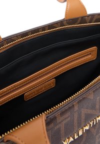 Indvendig i en brun mønstret taske med sort textilesyning, en brun lynlås lomme og et logo-tag. Guldfarvet lynlåsdetalje.