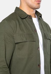 Olivově zelená košile-jacket vyrobená z pevné tkaniny, se zapínáním na knoflíky, dvěma náprsními kapsami a špičatým límcem.