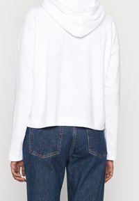 Felpa corta bianca con una consistenza morbida, dotata di cappuccio e maniche lunghe, indossata con jeans di denim blu scuro.