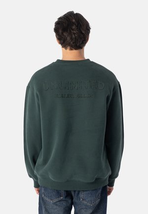 Persoon draagt een donkergroene trui met "UNLIMITED" en kleinere tekst op de achterkant, gecombineerd met een blauwe spijkerbroek, met de rug naar de camera.