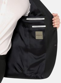Innenansicht eines schwarzen Blazers, mit glattem marineblauem Futter, zwei vertikalen Taschen, weißem Besatz und einem Etikett mit der Aufschrift "BENVENUTO."