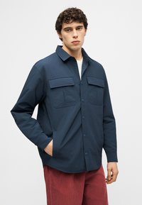 Ung mann med krøllete mørkt hår, iført en marineblå skjorte med knapper over en hvit topp og røde bukser, stående med én hånd i lomma.