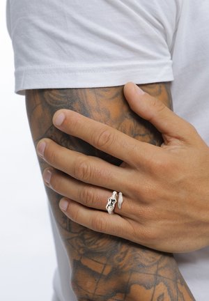 Hand mit silbernem künstlerischem Ring, der auf einem tätowierten Oberarm ruht, der von einem weißen kurzärmligen Hemd bedeckt ist.