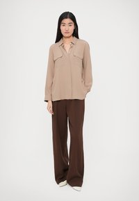 Beiges Hemd mit Knopfleiste und zwei Brusttaschen, kombiniert mit braunen, weit geschnittenen Hosen und weißen Schuhen mit spitzen Zehen, vor einem schlichten Hintergrund.