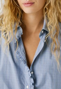 Gaiši zilas kvadrātu krekls ar viļņotiem malām ap pogu un apkakli. Iekļauj nelielas baltas pogas un brīvu piegriezumu.