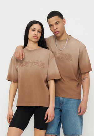 Νεαρός άνδρας και γυναίκα στέκονται δίπλα-δίπλα, φορώντας τα ίδια καφέ φαρδιά μπλουζάκια, η γυναίκα με μαύρα σορτς, ο άνδρας με τζιν σορτς και αλυσίδα στο λαιμό.