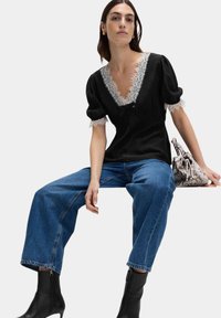 Schwarzes Oberteil mit Spitzenbesatz und puffärmel, kombiniert mit blauen weit geschnittenen Jeans. Das Model trägt schwarze Ankle-Boots und hält eine gemusterte Tasche.