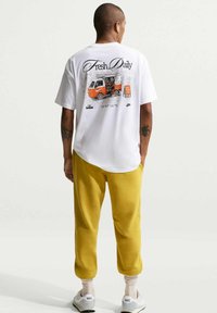 Homme portant un t-shirt blanc avec le graphisme "Fresh Daily" et une illustration orange de camion de nourriture, un pantalon moutarde, des baskets blanches Nike, et des écouteurs, debout.