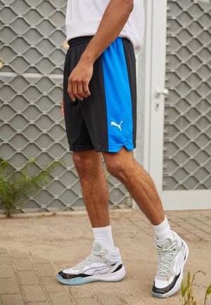 Homme portant un short de sport Puma noir et bleu avec des baskets blanches Puma et des chaussettes blanches, debout sur une surface pavée près d'une porte en métal.