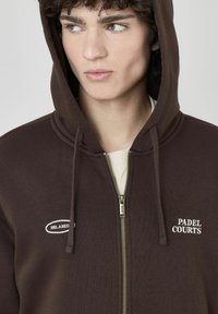 Hoodie zippé marron en tissu doux, avec une poche kangourou, des cordons de serrage et un texte brodé : « RELAXED » et « PADEL COURTS ». 