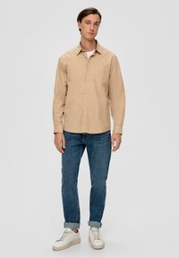 Beige Hemdbluse mit einer Tasche, kombiniert mit blauen Jeans und weißen Sneakern. Baumwollmaterial mit einer normalen Passform und langen Ärmeln.