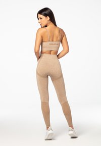 Beige sporta krūšturis un legingi, izgatavoti no elastīga auduma ar mīkstu tekstūru. Krūšturim ir aizmugurējais logo; legingiem ir tīkla akcenti.