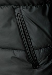 Detalle de chaqueta puff negra con cierre de cremallera visible, tela texturizada con patrones acolchados y un acabado mate suave.