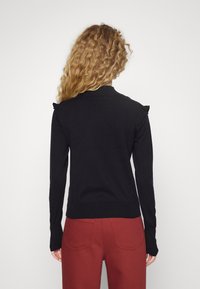 Kobieta o kręconych włosach, ubrana w dopasowany czarny top z długim rękawem i falbankami na ramionach oraz wysokim stanem pomarańczowe spodnie, pokazana od tyłu.